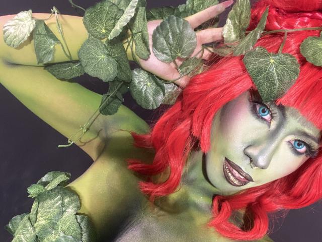 ScarletLuv with green skin, red hair, blue eyes, and dark lipstick, holding ivy leaves in her hair, showing her tits. She's looking at the camera with a sexy smile.