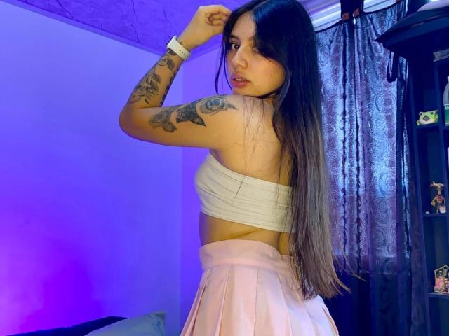 SophiaMarx with long brown hair, white tube top showing tits, pink pleated skirt, tattoos on right arm, looking back, purple-blue room light, black shelf with small items.