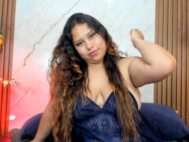 AnnyaMartinez with long wavy brown hair, wearing a blue lace dress, showing her tits, flexing her right arm, sitting on a dark blue couch.