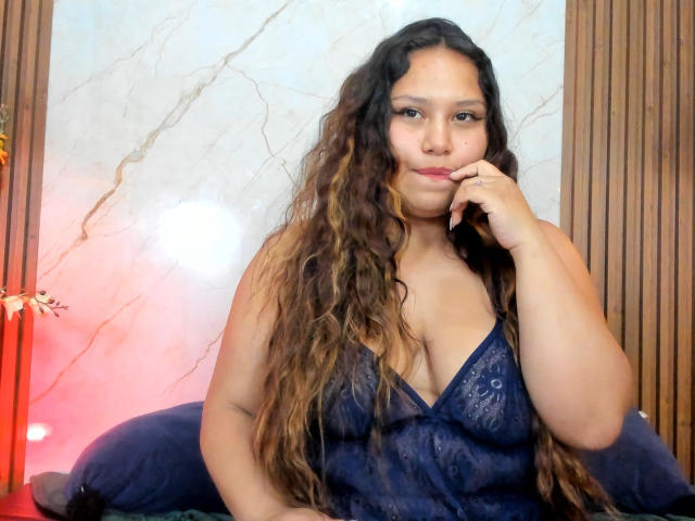 AnnyaMartinez with long wavy brown hair, wearing a blue lace dress, sitting on a bed, hand on chin, looking at camera, medium tits, soft smile, light brown skin.