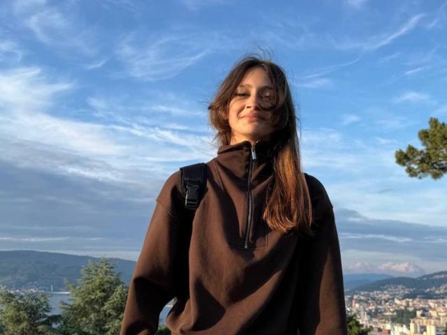 BernadineMelysa with brown hoodie, black backpack, and long brown hair, smiling under blue sky with wispy clouds. Her tits are small and her pussy is not visible.
