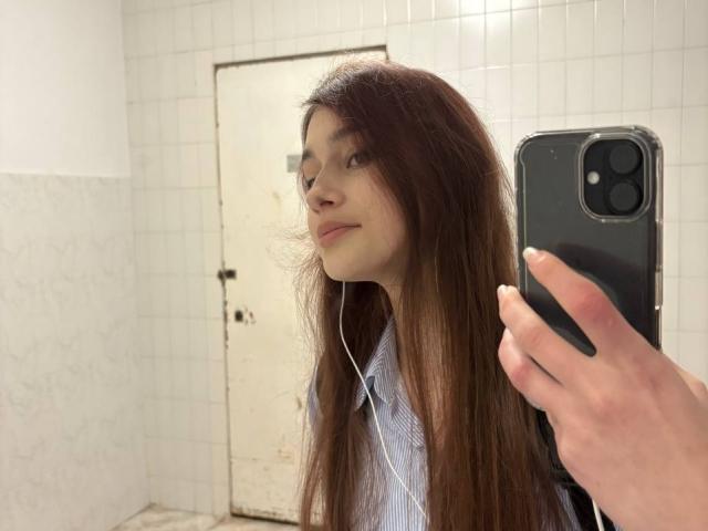 BernadineMelysa, com cabelos castanhos longos, fones de ouvido brancos e uma camisa azul listrada, tirando uma selfie com um iPhone preto, em um quarto com paredes de azulejos brancos, sorrindo levemente..