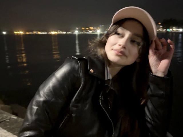 BernadineMelysa mit dunklem Haar, die einen rosa Hut und eine schwarze Lederjacke trägt, hält ihren Hut, Nacht am Wasser, Stadtlichter im Hintergrund, sanfter Gesichtsausdruck, leicht geneigter Kopf..