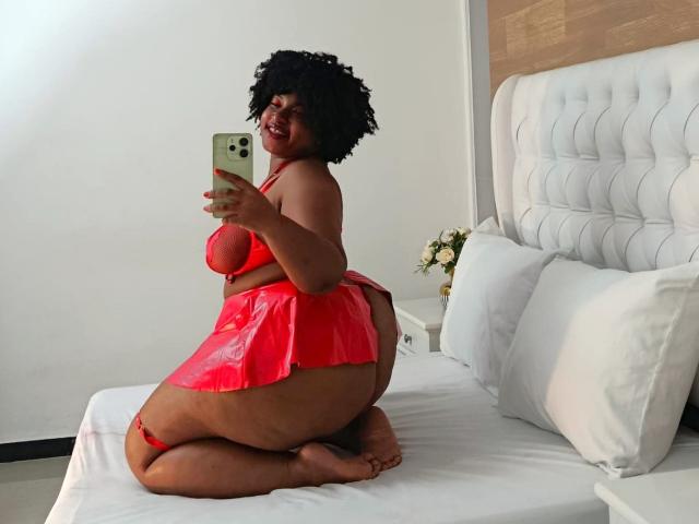 ViaraMoon with curly black hair, red latex lingerie, and a red thong, kneeling on a bed, showing her big ass and small tits, taking a selfie with a green phone.