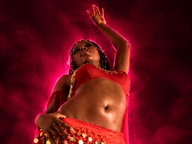 CatalinaSanA with dark skin, wearing a red bra and belly dancer outfit, raised arm, braided hair, glowing red background, midriff exposed, hand on hip, confident pose.