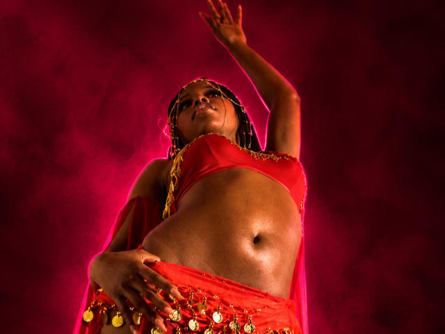 CatalinaSanA with dark skin, wearing a red bra and belly dancer outfit, raised arm, hand on hip, red smoke, gold jewelry, braided hair, looking down, glowing from below.