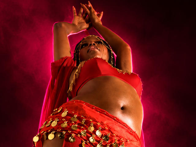 CatalinaSanA with dark skin, braided hair, red top, and red skirt with gold coins, arms raised, glowing pink background, showing her toned midriff and small tits.