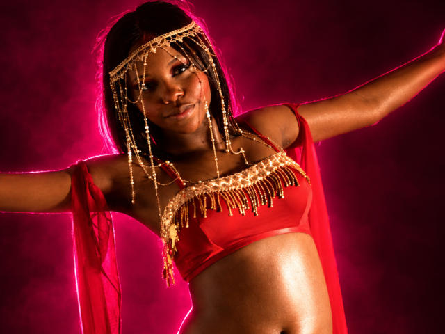 CatalinaSanA with dark skin, wearing a red top, gold chain headpiece, and red sheer sleeves, arms out, glowing pink background, shiny skin, small tits, confident smile.