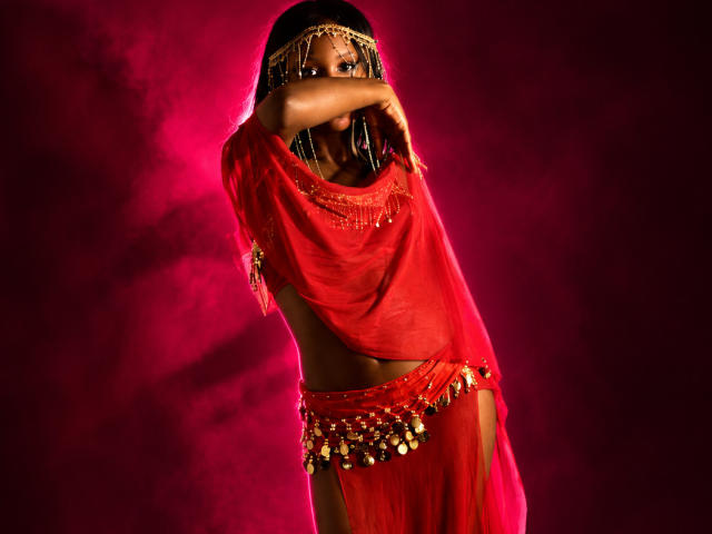 CatalinaSanA with dark skin, wearing a red sheer top and matching bottom with gold chains, arms covering her face, braided hair, red background, and a seductive pose.