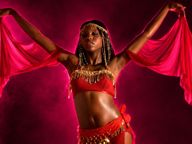 CatalinaSanA with dark skin, wearing a red bra and bottom with gold chains, red sheer sleeves, and a headpiece, holding them up, glowing pink background.