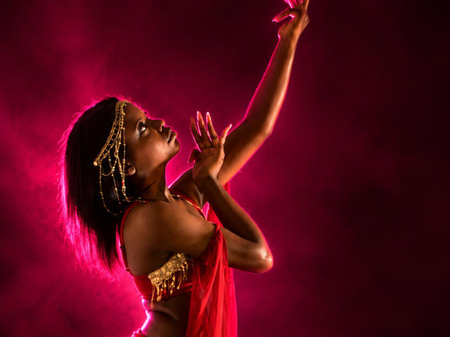CatalinaSanA with dark skin, wearing a red dress and gold headpiece, raises arms, showing off small tits and a toned ass against a pink-lit background.