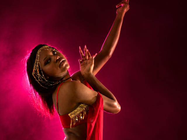 CatalinaSanA with dark skin, red dress, gold headpiece, and arm raised, looking back with a sultry expression. Her tits are visible, and she has a sexy, confident look.