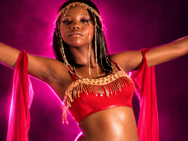 CatalinaSanA with dark skin, wearing a red bra and gold chain headpiece, arms out, red sheer sleeves, glowing pink background, showing her shiny tits and confident face.