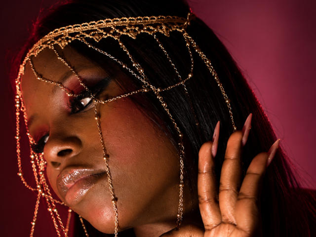 CatalinaSanA's face, dark skin, glossy lips, red eyeshadow, black hair, gold chain headpiece, long pink nails, right hand touching head, red background, serious expression, sexy.