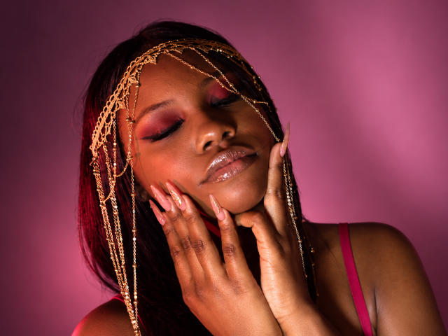 CatalinaSanA with glossy brown skin, pink eyeshadow, and gold chain headpiece, touching her shiny lips and hands with pink nails, wearing a pink top, against a pink background.