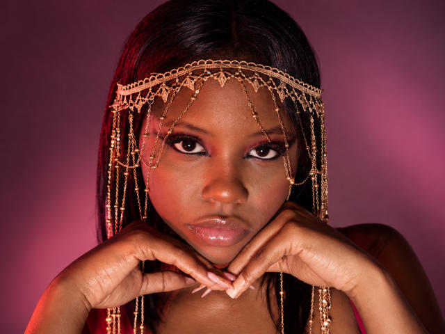 CatalinaSanA with dark skin, wearing a gold chain headpiece, has hands near her face, looking directly at the camera, pink and red lighting, glossy lips, and long nails.