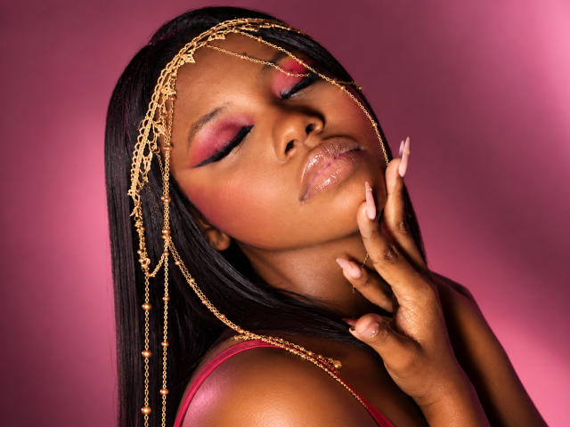 CatalinaSanA with dark skin, closed eyes, pink eyeshadow, glossy lips, and gold chain headpiece, touching her chin, wearing a red top, against a pink background.