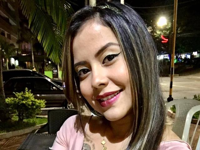 ElectraRoss with medium brown skin, black and blonde highlights, wearing pink off-shoulder top, smiling, pink lipstick, winged eyeliner, and necklace, standing outdoors at night.