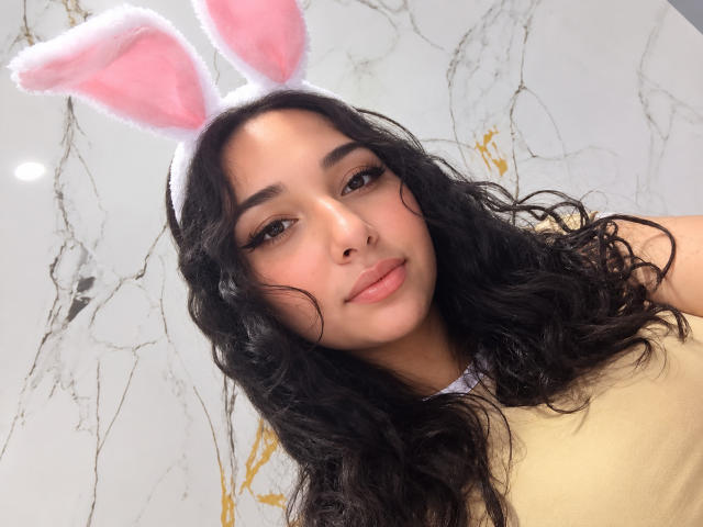 EleanorLamberth has wavy black hair, wearing pink and white fluffy bunny ears, with a cute smile and brown eyes, in a yellow top. Her ass is not visible.