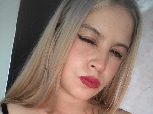 Melani Becker tiene el cabello rubio, guiña un ojo y hace pucheros con lápiz labial rojo. Sus pestañas son largas y lleva una blusa negra..