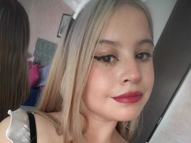 Melani Becker tiene el cabello rubio, lápiz labial rojo y delineador de ojos negro, sonríe con una cara linda, y lleva una blusa negra y blanca, mostrando un reflejo en el espejo..