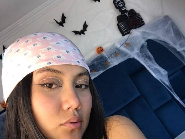Catalehia69 has light skin, long brown hair, wearing a pink floral beanie, and has glittery eye makeup, with a pouty expression, on a blue velvet headboard.