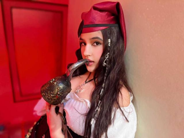IrisBlair with long black hair, wearing a red hat and white off-shoulder top, sucking on an ornate silver pipe, holding it with her right hand.