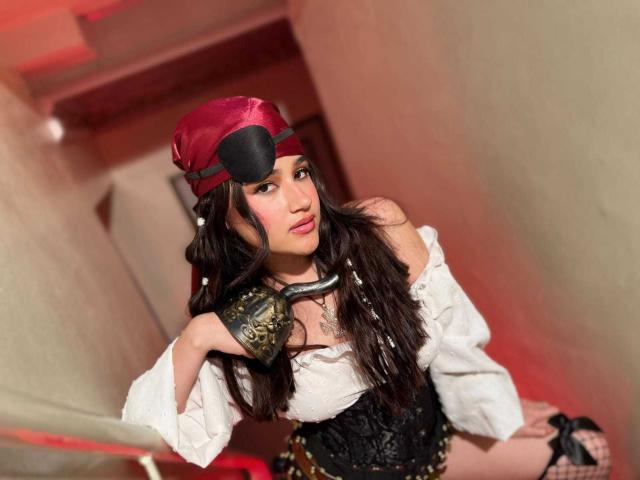 IrisBlair, with long brown hair, wearing a red hat, white off-shoulder shirt, black corset, and gold arm cuff, leaning against a staircase, showing her ass.