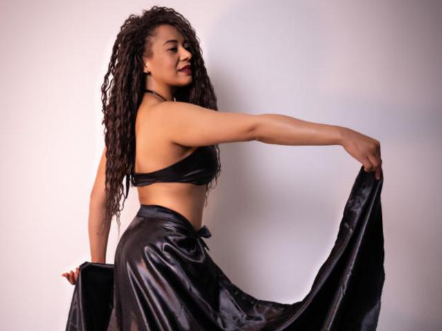 KenyaKarath with curly black hair, wearing a black bra and shiny black skirt, holding skirt, showing toned arms and small, firm tits, side profile, confident expression.