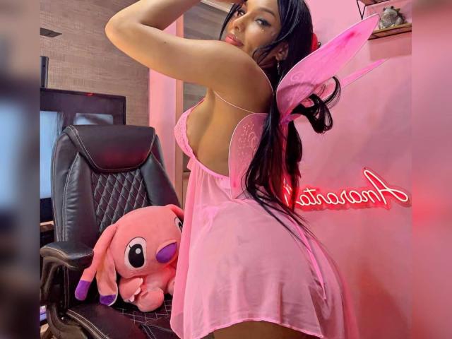 Amaranta69s in pink sheer lingerie, back to camera, showing ass, wearing pink fairy wings, long black hair, sitting on black chair with pink Plumpsy toy. Neon sign.