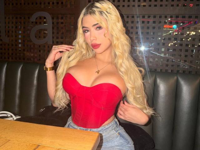 ChristieGarci with long blonde hair, red strapless top showing big boobs, denim shorts, gold bracelet, and red lipstick, sitting in a dark booth. Glowing light in background.