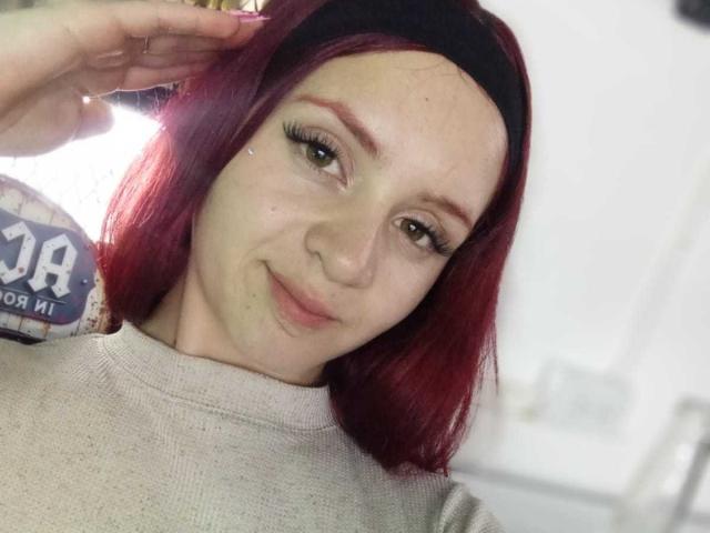 LesleyGray has red hair, green eyes, and a slight smile. She's wearing a black headband and a gray sweater. She's touching her hair with one hand.