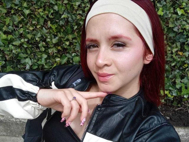 LesleyGray with red hair, wearing a beige headband, black and white jacket, making a "shhh" gesture with pink nails, has a nose ring and subtle makeup.