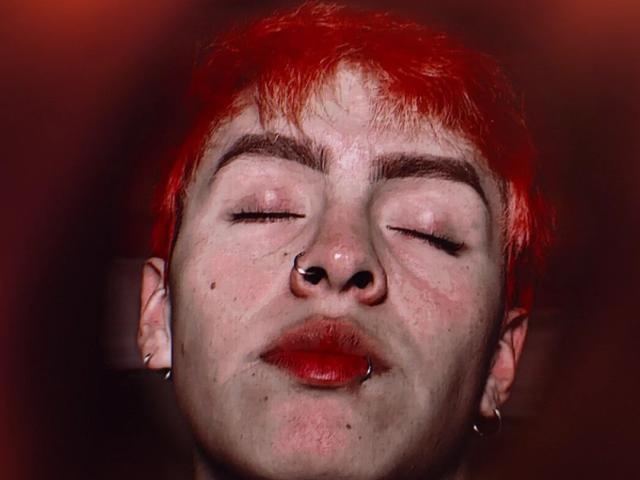 Babypeach07 ha i capelli rossi, gli occhi chiusi, un anello al naso e dei piercing in entrambe le orecchie, ed indossa un rossetto rosso. Il suo viso è al centro dell'immagine, con uno sfondo scuro..