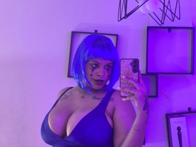 TeylorJenkins with blue bob, dark lipstick, and deep cleavage in a blue dress, holding a phone with a glittery case, purple lighting, tattoos on chest and hand.