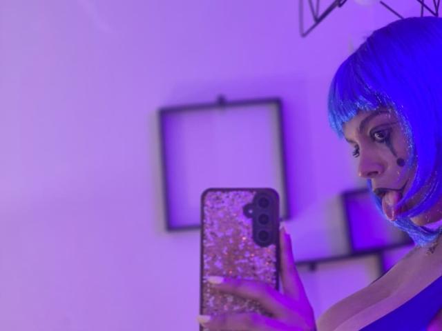TeylorJenkins with blue bob, makeup, and glitter phone case, taking a selfie, purple lighting, tongue out, looking at phone, holding it in her hand, visible cheek mark.