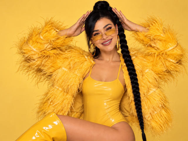 PaulinaValley with yellow latex bodysuit, yellow feather jacket, yellow thigh-high boots, long black braid, yellow glasses, smiling, hands in hair, yellow background, sexy, confident.