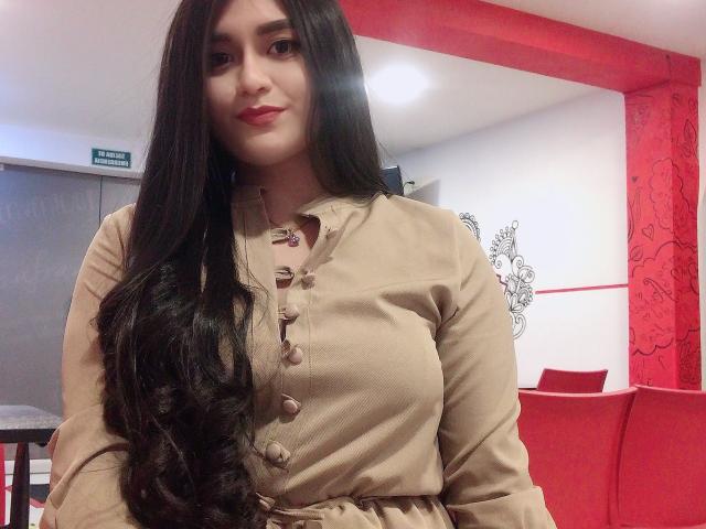 MiaDuboisX has long, wavy black hair, wearing a beige button-up dress, with buttoned chest, showing her tits slightly. She's in a red and white room, smiling.