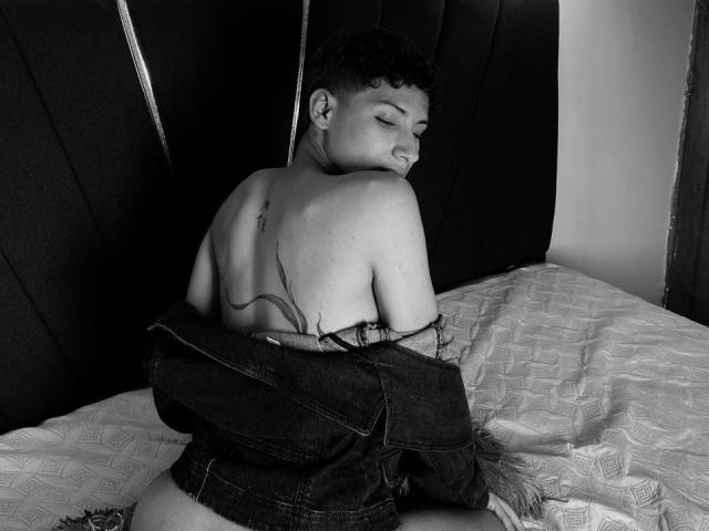 LuiisPey, topless with a denim jacket, showing a tattoo on their back, sitting on a bed, looking over their shoulder, with a slight smile, in a black-and-white photo.