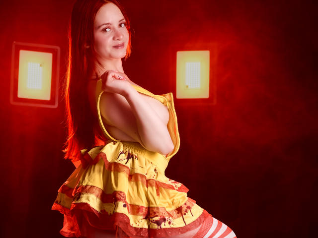 LesleyGray with red hair, yellow ruffled dress, and striped red-and-white stockings, smiling, holding dress strap, red lights in background, soft lighting, visible tits, cute, sexy.
