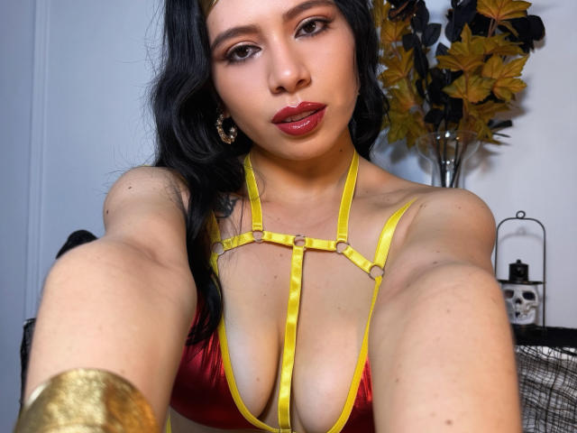 LindaMancini with dark hair, red lipstick, yellow harness over her tits, gold bracelet, and earrings, taking a selfie, looking at the camera, in a brightly lit room.