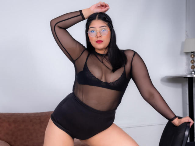 AlexaKiim with black sheer long-sleeve top, black lace bra, and high-waisted black shorts, posing with arm raised, wearing glasses, showing her tits and pussy through the top.