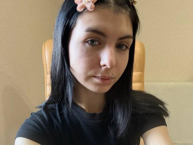 NobukoGraue, avec ses cheveux noirs, une pince à fleurs rose, des piercings au nez, un t-shirt noir, assise sur une chaise beige, peau pâle, yeux verts, lèvres légèrement pincées et une expression mignonne et sérieuse..