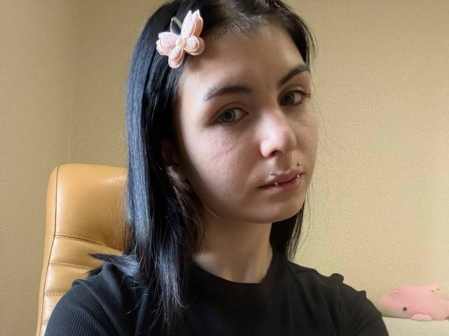 NobukoGraue a les cheveux noirs, une pince à cheveux en forme de papillon rose, un anneau au nez et un anneau aux lèvres. Elle porte une chemise noire et est assise sur un fauteuil en cuir beige..