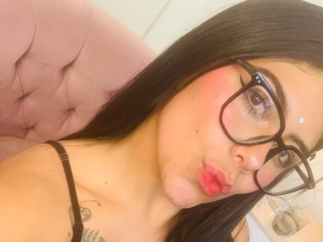 CatalinaReiyna with black hair, pink pout, and red lipstick, wearing big glasses, black strap, and a small tattoo on her shoulder, sitting on a pink tufted chair.