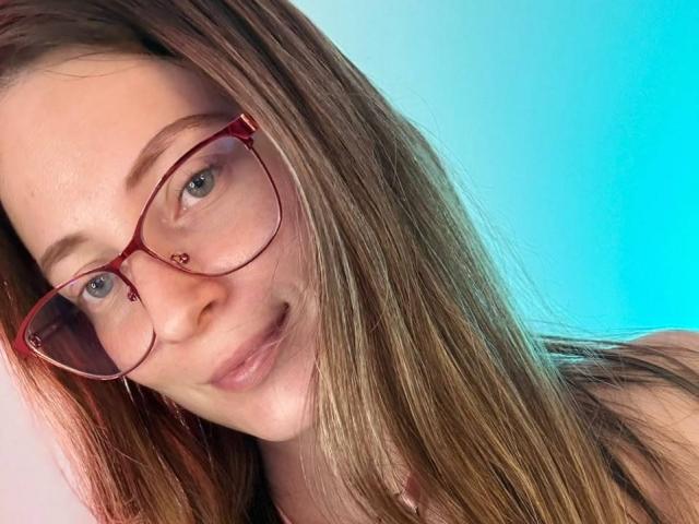 AmberWoody with red glasses, nose piercing, and long brown hair, smiling slightly, showing her cute face and green eyes, in a selfie with a blue background.