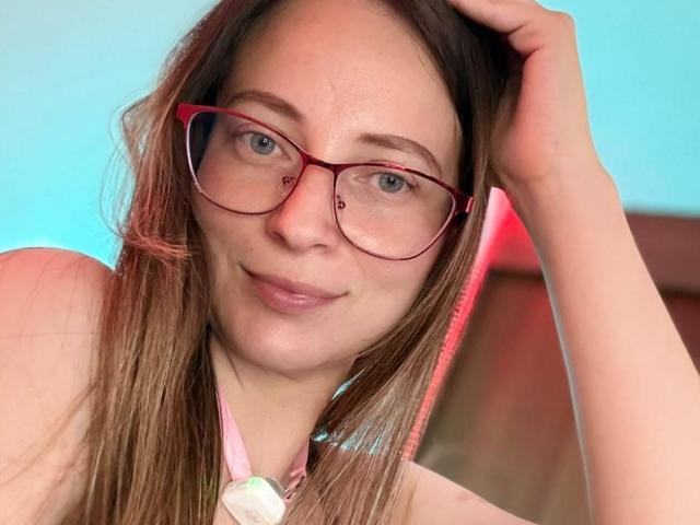 AmberWoody with red glasses, smiling, holding her brown hair. She's wearing a pink halter top, showing her bare shoulders and subtle tits. Light blue background.