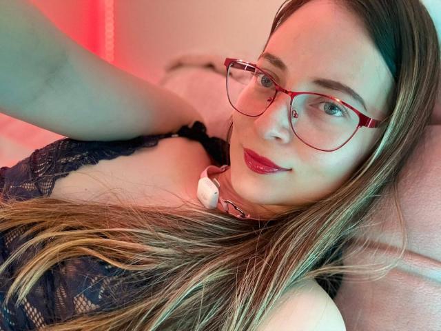 AmberWoody with red glasses, long brown hair, and red lipstick, wearing a black lace top and pink collar, smiling at the camera. Boobs visible, laying on pink pillow.