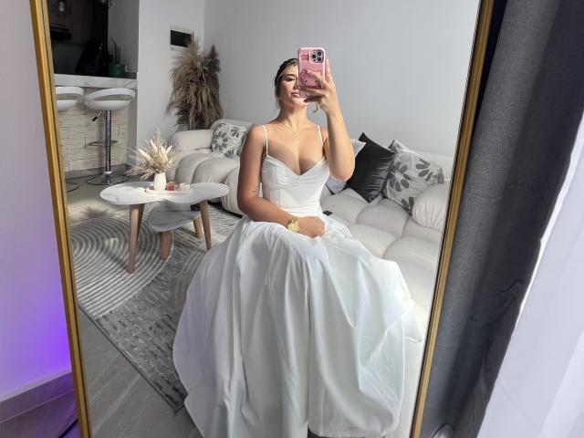 SashaPolet with tits in a white strapless dress, taking a mirror selfie, holding a pink phone, sitting on a white couch, smiling, in a modern, light-filled room.