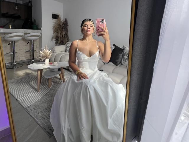 SashaPolet trägt ein weißes, trägerloses Brautkleid und hält ein rosa Handy. Im Spiegel ist ihr Spiegelbild zu sehen, das ihre Brüste und ihren Hintern zeigt. Sie trägt ein goldenes Armband an ihrem rechten Handgelenk..